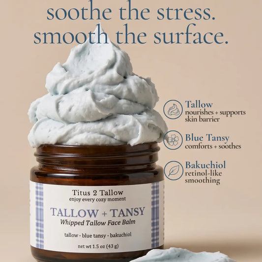 A jar of Tallow + Tansy Whipped Tallow Face Balm by Titus 2 Tallow, filled with pale blue whipped balm and topped with a large mound of the same balm. The amber glass jar sits on a warm beige background with marketing text and icons describing benefits.