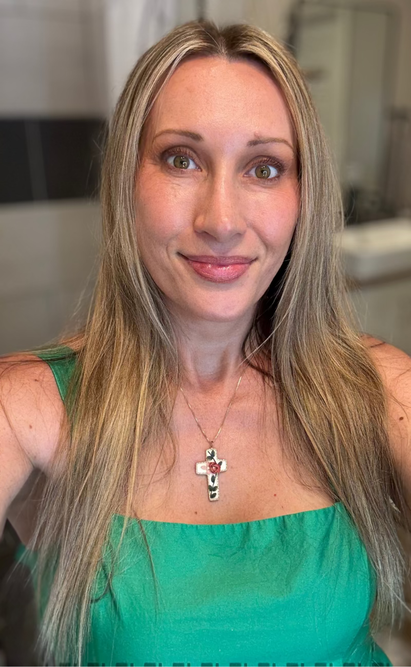 Woman with long blonde hair wearing a green top and a necklace with a cross pendant in a bathroom setting.